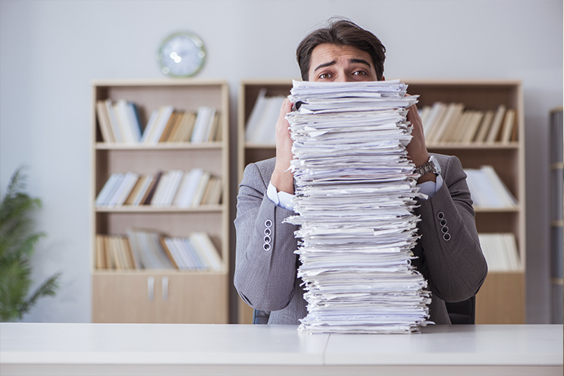 Um homem está no escritório e ele segura uma pliha de papeis, em cima da própria mesa, que chega até a altura de seus olhos. Ele tem um olhar de aflição porque não sabe como lidar com tanta burocracia.