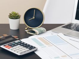 Em cima de uma mesa está um laptop, um relógio, um bolo de dinheiro, moedas, um pequeno vaso, calculadora e folhas impressas com tabelas coloridas. Parece se tratar de um ambiente doméstico em que a pessoa usa para trabalhar. A ideia é mostrar que o empresário não deve misturar as contas da pessoa física com a jurídica. O melhor a se fazer é cuidar do controle financeiro.