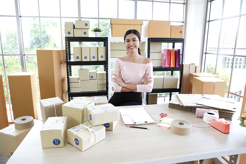 Uma mulher está numa das salas de sua empresa. Ela está sorrindo, atrás de uma grande mesa, onde há pacotes separados para entrega por encomenda. A ideia é mostrar que o microempresário pode exportar seu produtos.