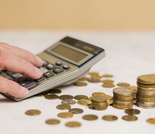 Calculadora sendo usada por um homem. Ao lado dela, há várias moedas espalhadas pela mesa. A ideia é mostrar que o momento atual é de fazer as contas para aproveitar as oportunidades de negócios a partir da nova taxa de juros.
