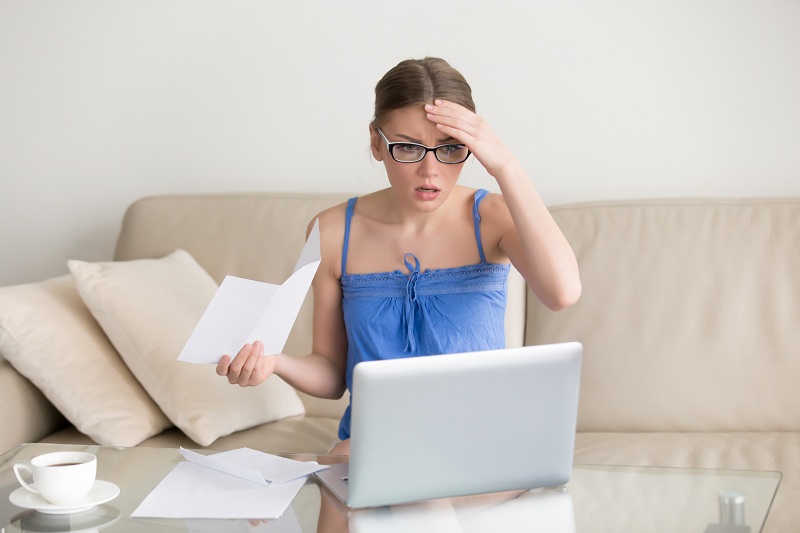 Mulher está sentada no sofá de casa, com seu laptop numa mesa de vidro que está na sua frente. Com a mão esquerda na testa, ela segura alguns papeis com a mão direita e faz cara de espanto ao olhar para a tela do computador. Ela deve estar olhando o extrato do banco e está assustada com a possível dívida que tem no cheque especial ou cartão de crédito.