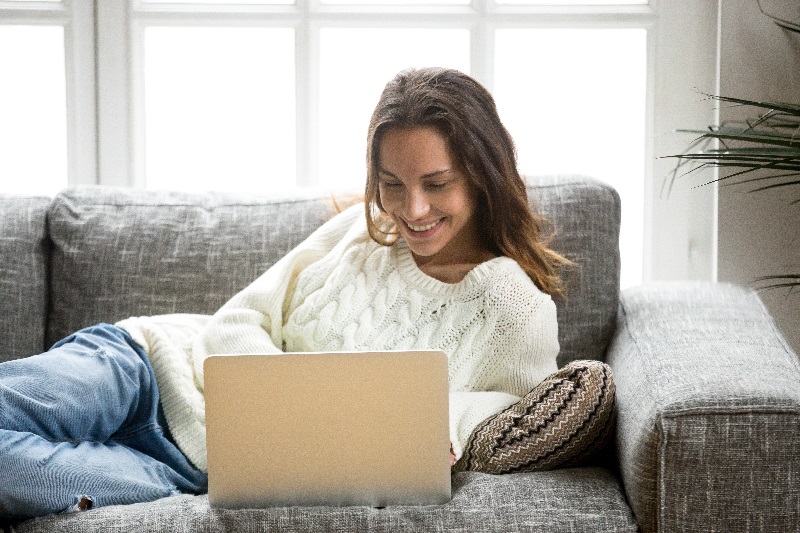 Mulher está deitada no sofá, lendo o blog em seu computador.