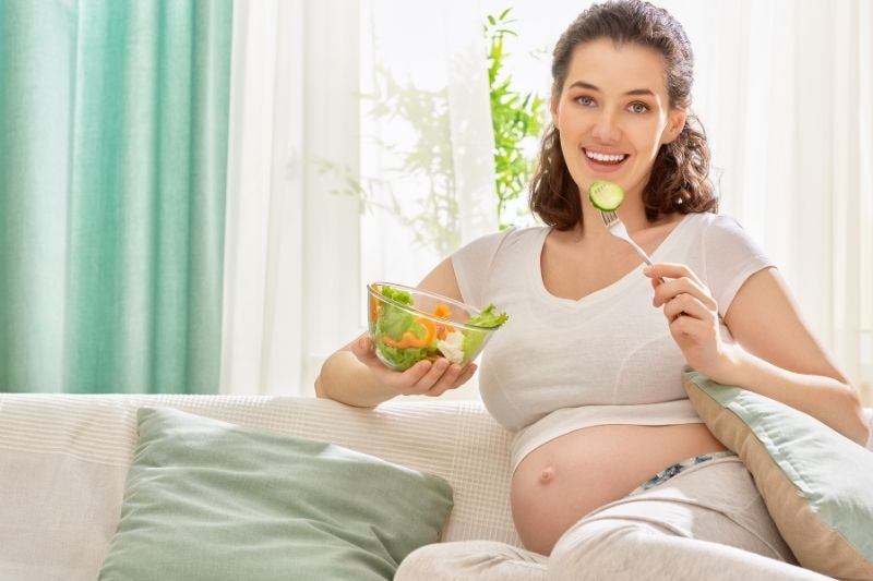 Mulher está sentada no sofá, comendo uma salada, para garantir a alimentação saudável.