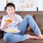 Menino está sentado no sofá e comendo hamburguer com batata frita, que é péssimo para alimentação infantil.