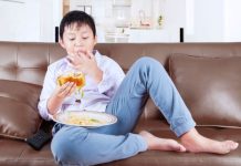 Menino está sentado no sofá e comendo hamburguer com batata frita, que é péssimo para alimentação infantil.