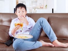 Menino está sentado no sofá e comendo hamburguer com batata frita, que é péssimo para alimentação infantil.