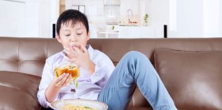 Menino está sentado no sofá e comendo hamburguer com batata frita, que é péssimo para alimentação infantil.