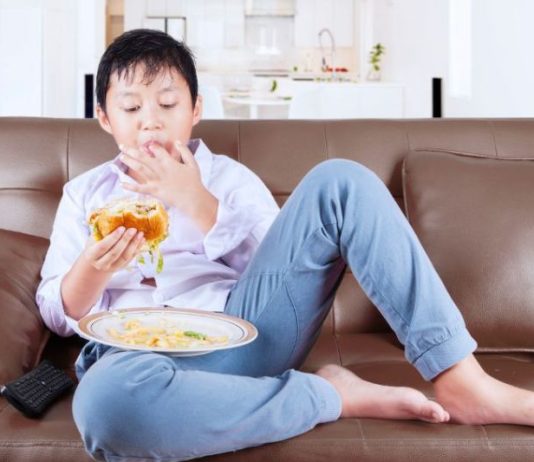 Menino está sentado no sofá e comendo hamburguer com batata frita, que é péssimo para alimentação infantil.